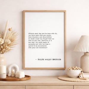 May include: A framed print with a quote by Ralph Waldo Emerson, displayed on a wooden surface with decorative items. The quote is in black text on a white background. A vase of dried flowers, candles, and a lamp are also visible.