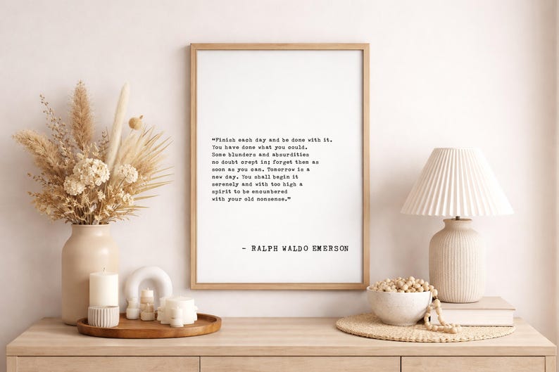 May include: A framed print with a quote by Ralph Waldo Emerson, displayed on a wooden surface with decorative items. The quote is in black text on a white background. A vase of dried flowers, candles, and a lamp are also visible.