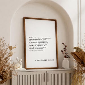 May include: A framed print with a quote by Ralph Waldo Emerson. The text is in black on a white background, set within a dark brown wooden frame. The quote reads, "Finish each day and be done with it..." The print is displayed on a white cabinet.