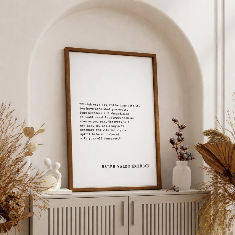 May include: A framed print with a quote by Ralph Waldo Emerson. The text is in black on a white background, set within a dark brown wooden frame. The quote reads, "Finish each day and be done with it..." The print is displayed on a white cabinet.