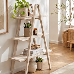 May include: A light-colored wooden A-frame shelving unit with three horizontal shelves. The shelves hold a potted green plant, a small picture frame, a succulent in a textured pot, a candle, a wooden chain link, two books, and a ceramic vase.