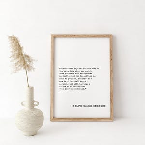 May include: A framed print with a quote by Ralph Waldo Emerson in black text on a white background. The frame is light brown. A decorative vase with a dried plant is to the left of the frame.