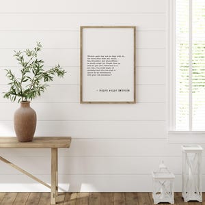 May include: A framed print with a quote by Ralph Waldo Emerson. The text is in black font on a white background, framed in a light brown wooden frame. The print hangs on a white shiplap wall. A wooden bench and a vase with greenery are in the foreground.