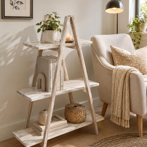 May include: A three-tier A-frame shelving unit made of light-colored wood with a distressed white finish. The triangular structure features three horizontal shelves that decrease in width toward the top, providing open storage for various decorative items.