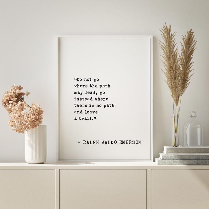 May include: A framed print with a white background features a quote by Ralph Waldo Emerson in black text: "Do not go where the path may lead, go instead where there is no path and leave a trail." Decorative elements include dried flowers and a vase.