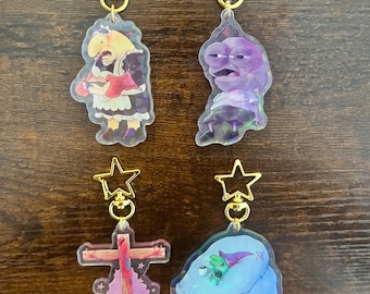 May include: Four acrylic keychains with gold star clasps. The keychains feature cartoon characters in various poses. One keychain shows a figure in a red and white dress. Another shows a purple frog-like character. The other two keychains show a cross and a green creature.