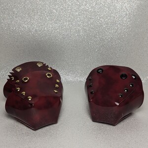 May include: Two burgundy leather wrist cuffs with gold-toned metal accents and black rivets. The cuffs are closed with a buckle.