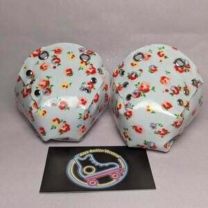 May include: Two white roller skate toe stops with a floral pattern in red, yellow, and blue. The toe stops have silver eyelets and rivets.
