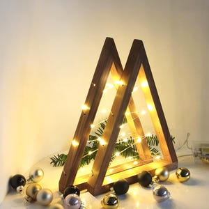 Op de afbeelding: Een driehoekig houten frame verlicht door een snoer met warmwitte LED-lampjes. Het frame is geplaatst over groene dennennaalden en wordt omringd door kleine, ronde kerstballen in de kleuren goud, zwart en zilver.