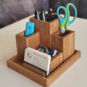 Könnte beinhalten: Ein Schreibtisch-Organizer aus Holz mit drei quadratischen Fächern unterschiedlicher Höhe auf einer quadratischen Basis. Er enthält schwarze Stifte, einen hellblauen Textmarker, eine grün-blaue Schere und Visitenkarten mit dem Text Hölzchen Stoffchen.
