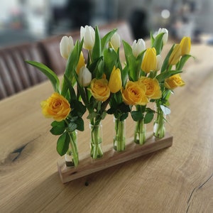 Peut inclure: Un vase à fleurs en bois avec six tubes en verre contient un bouquet de roses jaunes et de tulipes blanches. Le vase est posé sur une table en bois.