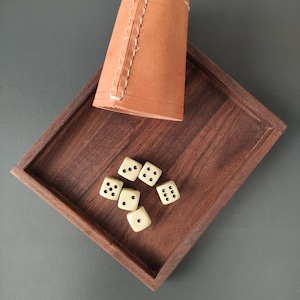 Op de afbeelding: Een houten dienblad met zes witte dobbelstenen en een bruine leren dobbelbeker. De dobbelstenen liggen verspreid op het dienblad.