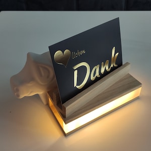 K&ouml;nnte beinhalten: Ein beleuchtetes Holzdisplay mit einer schwarzen Karte, auf der "Lieben Dank" mit einem goldenen Herzen und goldenen Buchstaben steht. Das Display wird von unten beleuchtet und erzeugt ein warmes Leuchten.