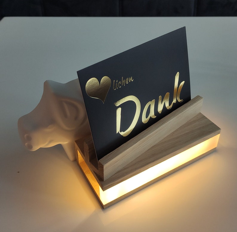 K&ouml;nnte beinhalten: Ein beleuchtetes Holzdisplay mit einer schwarzen Karte, auf der "Lieben Dank" mit einem goldenen Herzen und goldenen Buchstaben steht. Das Display wird von unten beleuchtet und erzeugt ein warmes Leuchten.