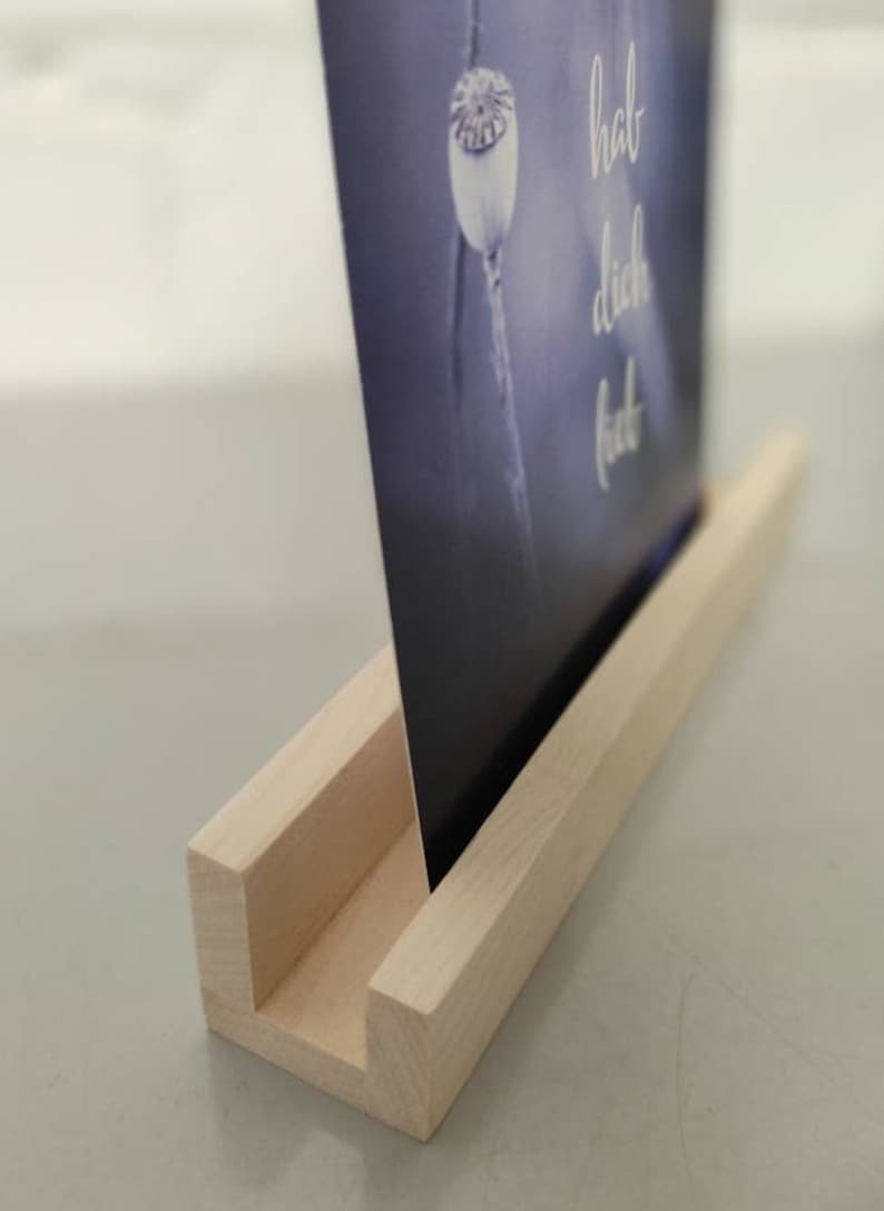 K&ouml;nnte beinhalten: Ein Holzbildst&auml;nder mit einer hellen Holzoptik. Der St&auml;nder hat einen Schlitz, um ein Bild oder eine Karte zu halten. Die Karte im St&auml;nder hat einen blauen Hintergrund mit einer wei&szlig;en Mohnkapsel und dem Text "hab dich lieb".