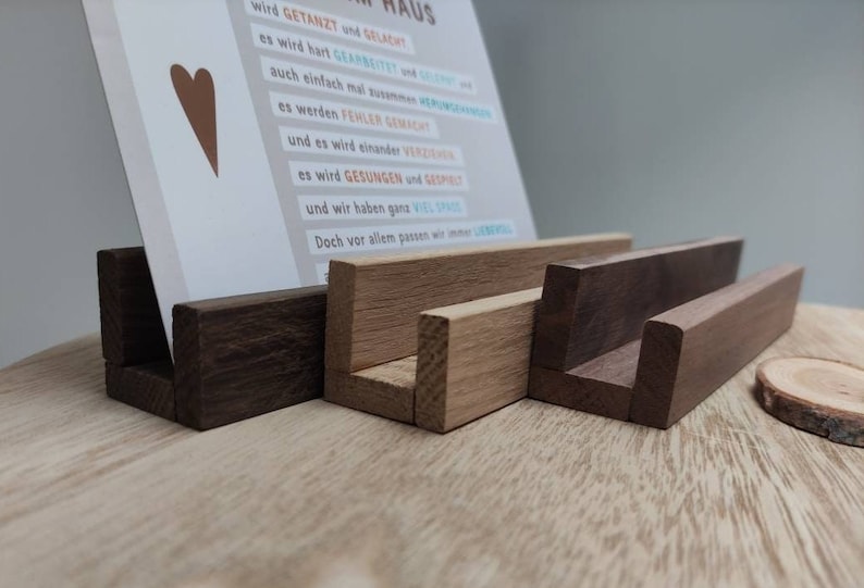 K&ouml;nnte beinhalten: Vier Kartenhalter aus Holz in verschiedenen Braunt&ouml;nen. Die Halter sind aus verschiedenen Holzarten gefertigt, darunter Nussbaum und Eiche. Die Halter sind in einer Reihe angeordnet, wobei der gr&ouml;&szlig;te Halter links und der kleinste Halter rechts ist. Die Halter sind so konzipiert, dass sie Karten oder andere kleine Gegenst&auml;nde aufrecht halten.