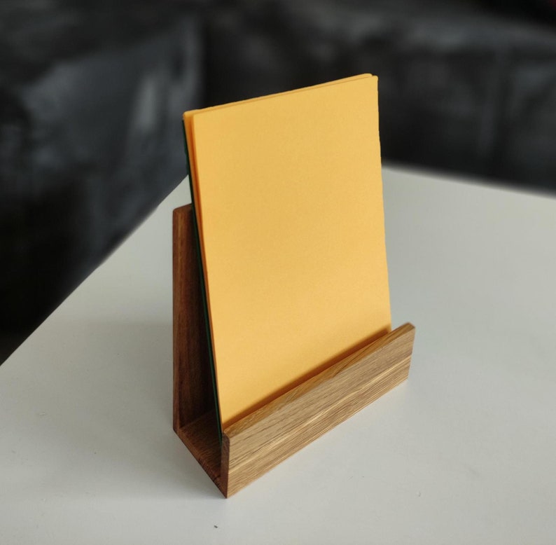 May include: A wooden stand with a light brown finish holds a stack of yellow paper. The stand has a simple design with a rectangular base and a vertical back.