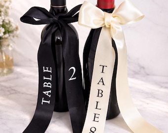 May include: Two wine bottles with black and cream-colored ribbons. The black ribbon reads "TABLE 2" in white, while the cream ribbon says "TABLE 8". The bottles are decorated for a wedding or event.