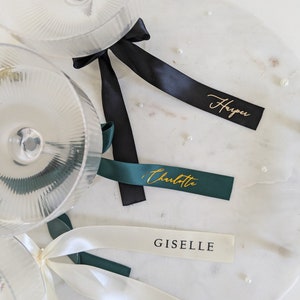May include: Three clear ribbed glass coupes with satin ribbons tied around the stems. One black ribbon features the gold script name Harper, one dark green ribbon features the gold script name Charlotte, and one white ribbon features the name GISELLE.