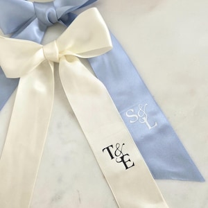 May include: Two satin ribbon bows, one ivory and one light blue, with long tails. The ivory ribbon has the initials "T&E" printed in black. The light blue ribbon has the initials "S&L" embroidered in white.
