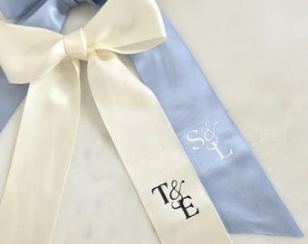 May include: Two satin ribbon bows, one ivory and one light blue, with long tails. The ivory ribbon has the initials "T&E" printed in black. The light blue ribbon has the initials "S&L" embroidered in white.