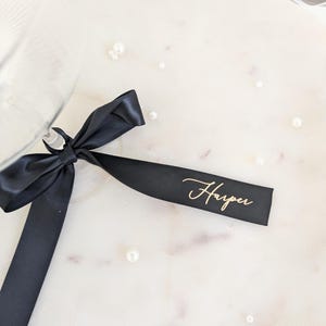 May include: A black satin ribbon tied in a bow around the stem of a glass. The ribbon features the name Harper printed in a gold cursive script font. The ribbon is positioned against a light surface scattered with small white pearls.