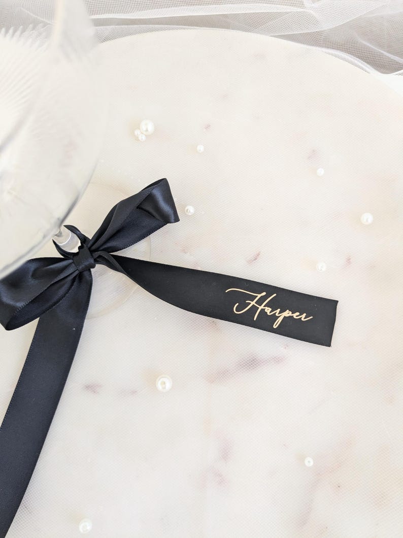 May include: A black satin ribbon tied in a bow around the stem of a glass. The ribbon features the name Harper printed in a gold cursive script font. The ribbon is positioned against a light surface scattered with small white pearls.