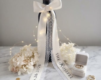 May include: A dark wine bottle adorned with a white satin bow and long ribbons featuring lace trim. The ribbons are printed with the text 06.26.2026 and THE RUSSELL. A string of fairy lights, two white hydrangea blooms, a ring box, and a gold key.