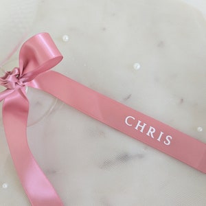 May include: A dusty rose satin ribbon tied in a bow, featuring the name CHRIS printed in white capital letters on the fabric. The smooth, reflective surface of the ribbon contrasts with the soft, sheer, off-white tulle fabric underneath.