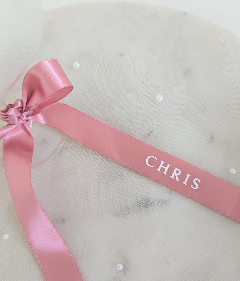 May include: A dusty rose satin ribbon tied in a bow, featuring the name CHRIS printed in white capital letters on the fabric. The smooth, reflective surface of the ribbon contrasts with the soft, sheer, off-white tulle fabric underneath.