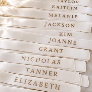 May include: A collection of white satin ribbons, each featuring a name printed in gold capital letters. The names listed are Victoria, Taylor, Kaitlin, Melanie, Jackson, Kim, Joanne, Grant, Nicholas, Tanner, Elizabeth, and Devin, arranged in rows.