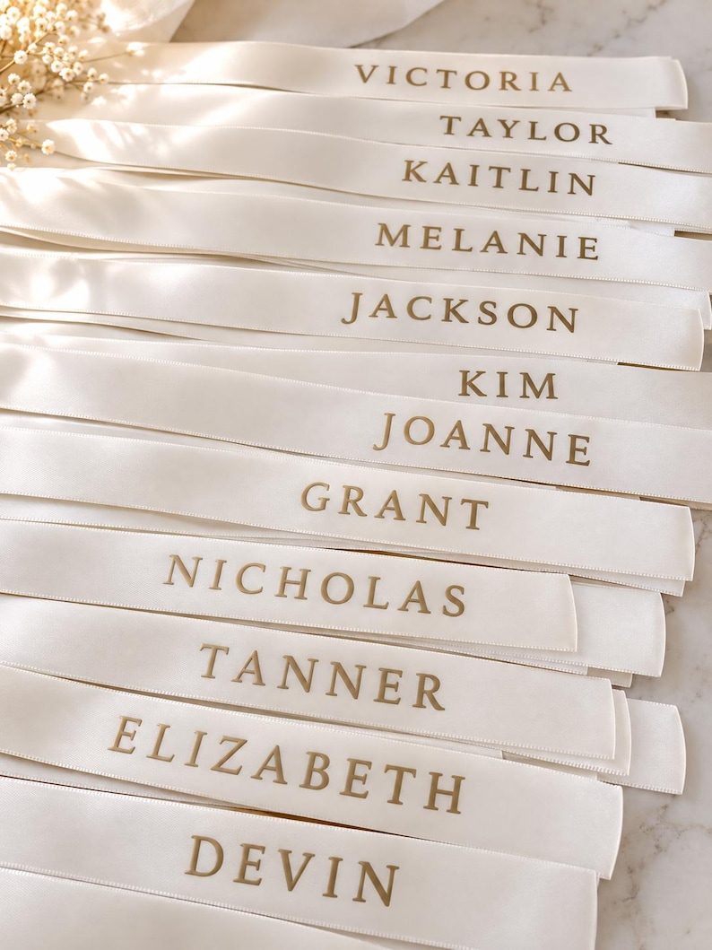May include: A collection of white satin ribbons, each featuring a name printed in gold capital letters. The names listed are Victoria, Taylor, Kaitlin, Melanie, Jackson, Kim, Joanne, Grant, Nicholas, Tanner, Elizabeth, and Devin, arranged in rows.