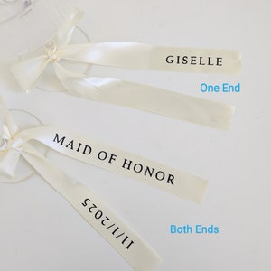 May include: Two cream-colored satin ribbons tied to glass stems. The top ribbon features the text GISELLE on one end. The bottom ribbon features the text MAID OF HONOR and 11/1/2025 on both ends. The text One End and Both Ends is visible.