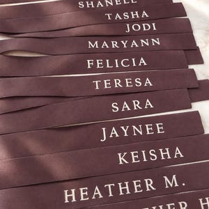 May include: A collection of dark brown satin ribbons, each featuring a name printed in white capital letters. The names include Jeanine, Shanell, Tasha, Jodi, Maryann, Felicia, Teresa, Sara, Jaynee, Keisha, Heather M., and Heather H. arranged in rows.