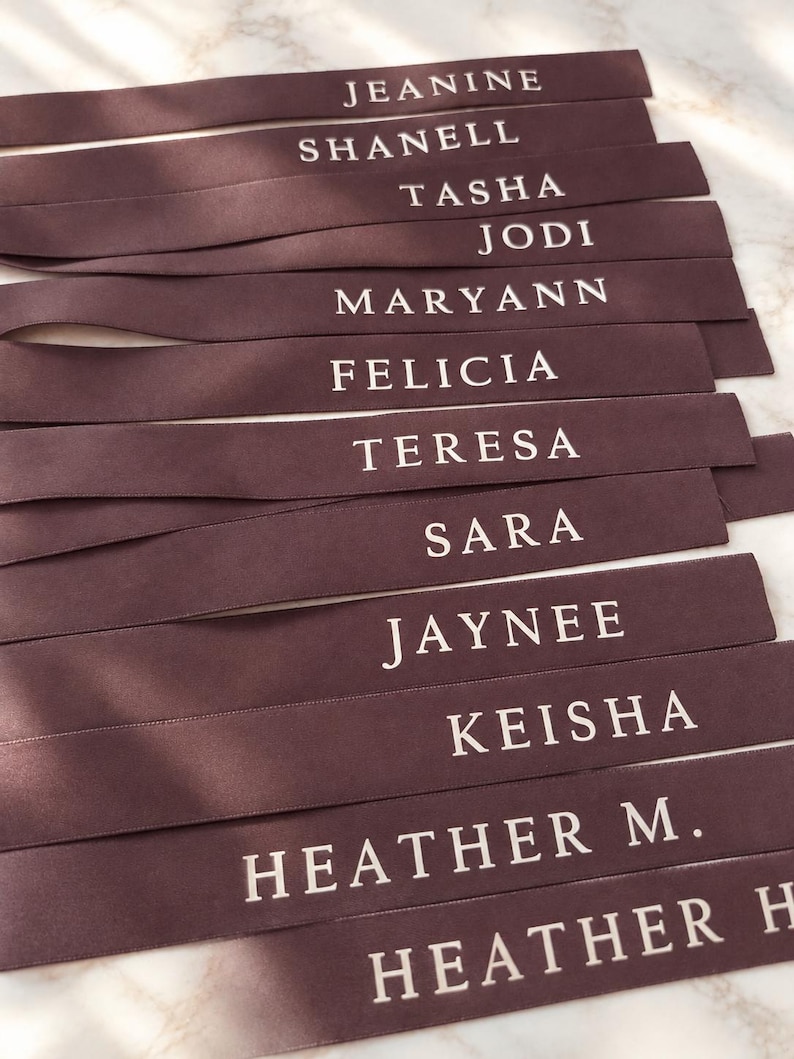 May include: A collection of dark brown satin ribbons, each featuring a name printed in white capital letters. The names include Jeanine, Shanell, Tasha, Jodi, Maryann, Felicia, Teresa, Sara, Jaynee, Keisha, Heather M., and Heather H. arranged in rows.