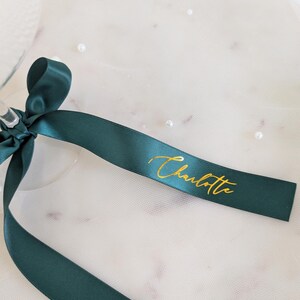 May include: A dark green satin ribbon tied in a bow, featuring the name Charlotte printed in metallic gold cursive script. The ribbon is smooth and reflective, with the gold lettering clearly visible against the deep green fabric surface.