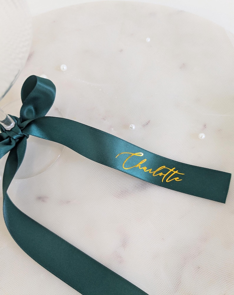 May include: A dark green satin ribbon tied in a bow, featuring the name Charlotte printed in metallic gold cursive script. The ribbon is smooth and reflective, with the gold lettering clearly visible against the deep green fabric surface.