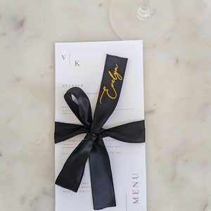 May include: A white rectangular menu card featuring the letters VK at the top and the word MENU vertically on the right side. A black satin ribbon is tied around the center, with the name Evelyn written in gold cursive script on the ribbon.
