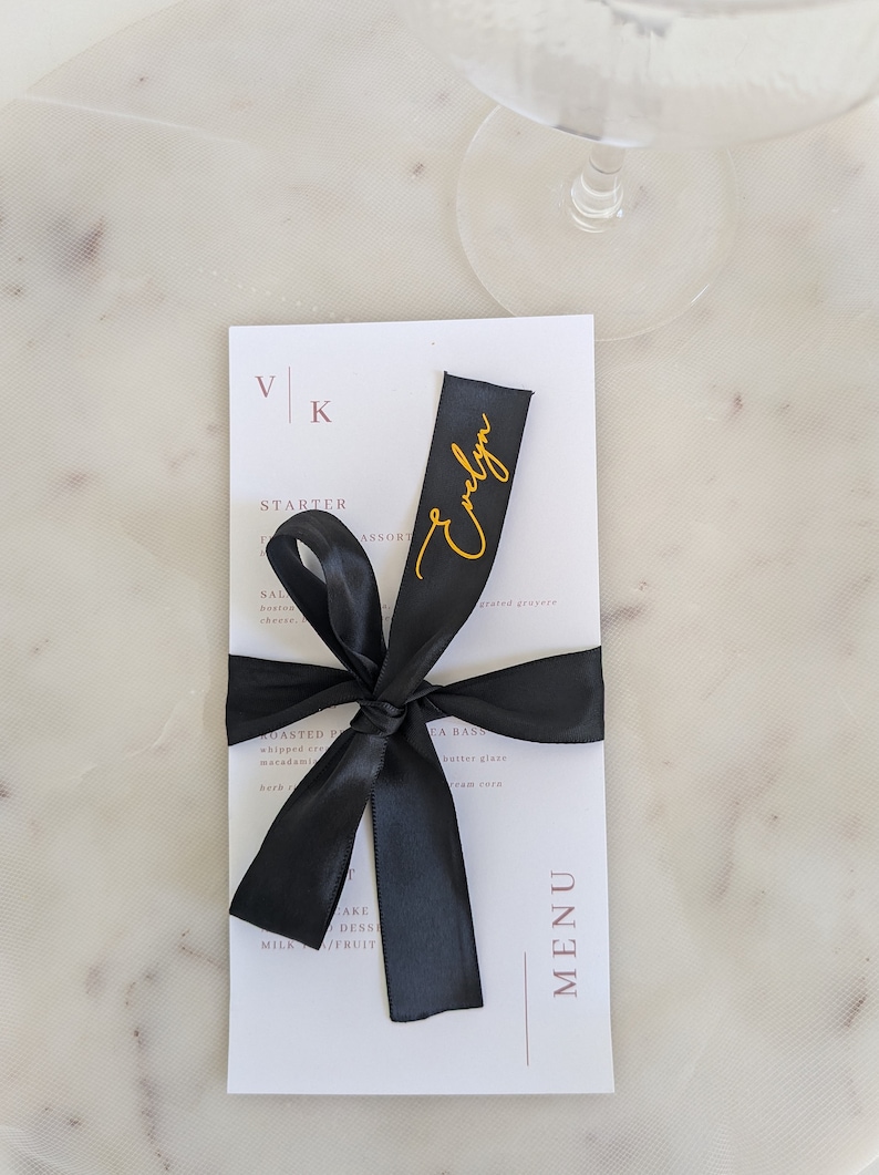 May include: A white rectangular menu card featuring the letters VK at the top and the word MENU vertically on the right side. A black satin ribbon is tied around the center, with the name Evelyn written in gold cursive script on the ribbon.