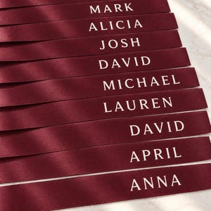 May include: A stack of ten burgundy satin ribbons, each featuring a white printed name. The names listed from top to bottom are PHILLIP, MARK, ALICIA, JOSH, DAVID, MICHAEL, LAUREN, DAVID, APRIL, and ANNA. The ribbons have a smooth, reflective finish.