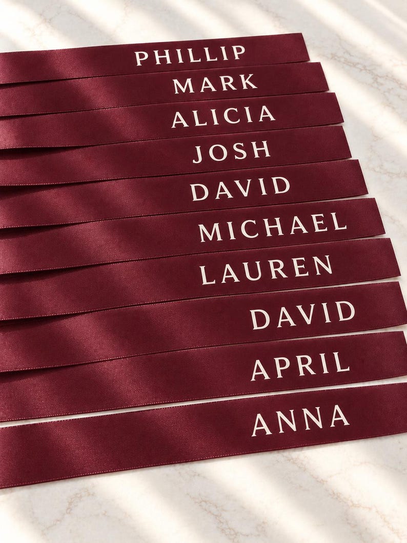 May include: A stack of ten burgundy satin ribbons, each featuring a white printed name. The names listed from top to bottom are PHILLIP, MARK, ALICIA, JOSH, DAVID, MICHAEL, LAUREN, DAVID, APRIL, and ANNA. The ribbons have a smooth, reflective finish.