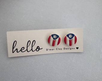 Puede incluir: Un par de pendientes redondos con el diseño de la bandera puertorriqueña. Los pendientes tienen una base blanca con rayas rojas y una sección azul con una estrella blanca. Se exhiben en una tarjeta con "hello" y "D'mar Clay Designs".