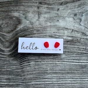 May include: A pair of bright red, textured raspberry-shaped stud earrings attached to a white rectangular display card. The card features the word hello in black script, the text D'mar Clay Designs, and a small black heart symbol.