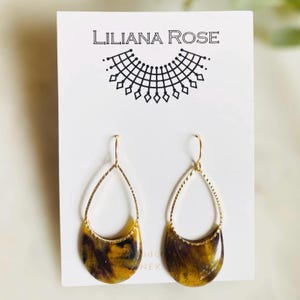 Könnte beinhalten: Goldene Tropfenohrringe mit einem marmorierten Design in Gelb, Schwarz und Braun. Die Ohrringe befinden sich auf einer weißen Karte mit dem aufgedruckten Text "Liliana Rose".