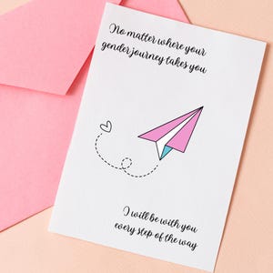 May include: A white greeting card with the text "No matter where your gender journey takes you" and "I will be with you every step of the way." The card features a pink, white, and blue paper airplane illustration and a small heart.