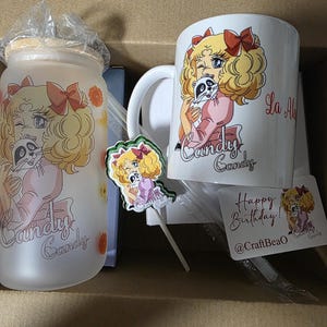 Peut inclure: Un gobelet en verre dépoli avec couvercle en bambou, une tasse en céramique blanche, une petite figurine sur bâtonnet et une carte d'anniversaire. Chaque objet présente une illustration de fille blonde aux nœuds rouges tenant un raton laveur.
