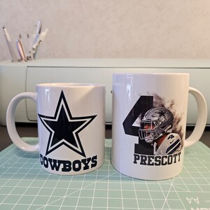 May include: Two white ceramic mugs with a Dallas Cowboys theme. One mug features a blue star with the word "COWBOYS" underneath. The other mug has a photo of a football player with the number "4" and the name "PRESCOTT" underneath.