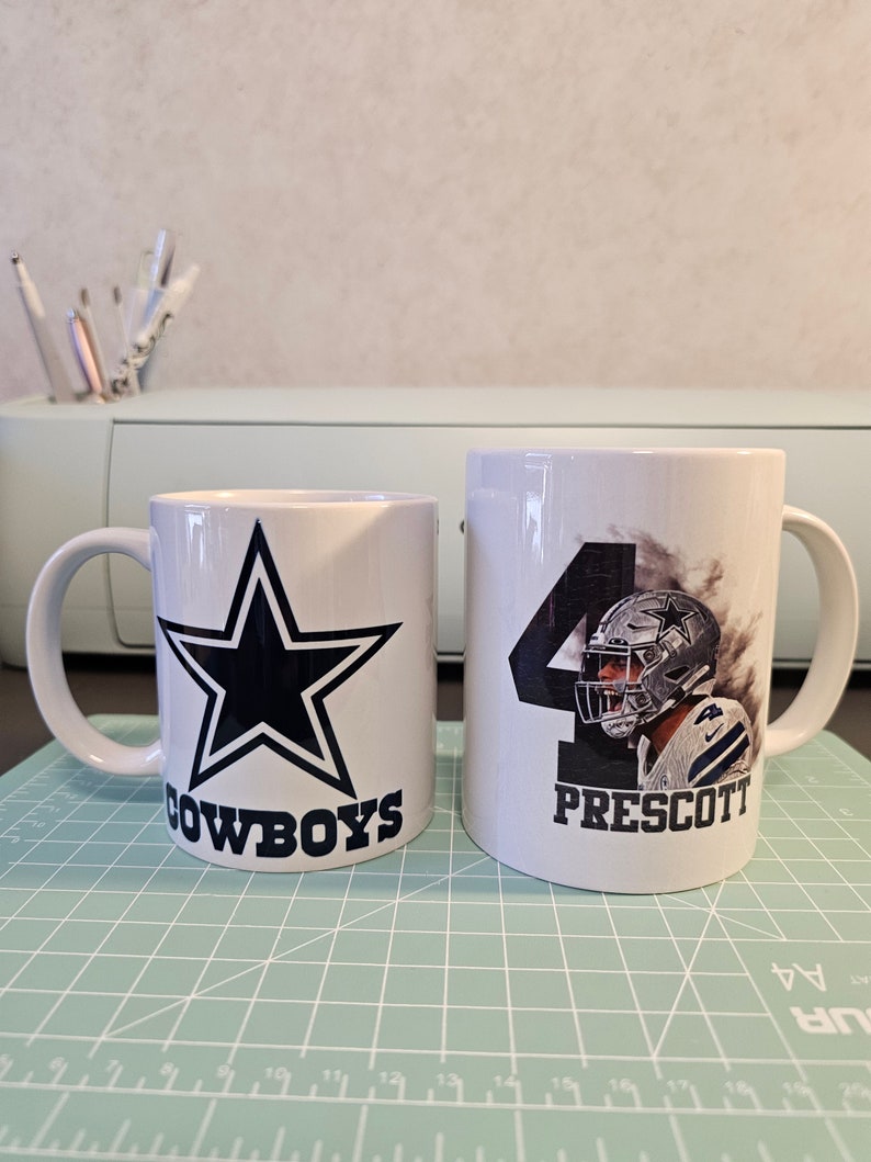 May include: Two white ceramic mugs with a Dallas Cowboys theme. One mug features a blue star with the word "COWBOYS" underneath. The other mug has a photo of a football player with the number "4" and the name "PRESCOTT" underneath.