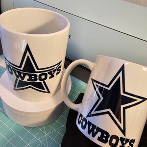 May include: Two white ceramic mugs with black and blue Dallas Cowboys logos. The mugs are on a green cutting mat.