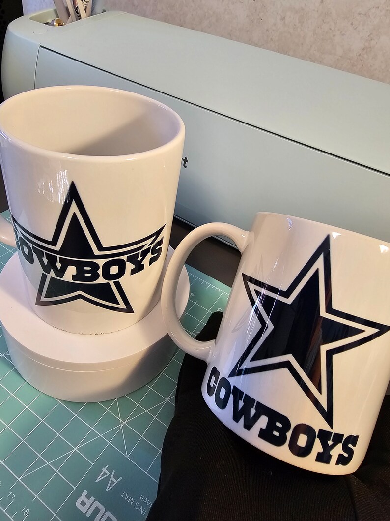 May include: Two white ceramic mugs with black and blue Dallas Cowboys logos. The mugs are on a green cutting mat.